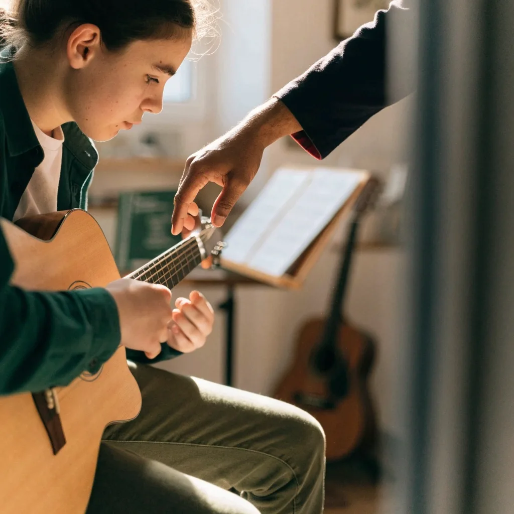 A tutor adjusting a student's fretting hand on an acoustic guitar during a lesson in a small teaching room