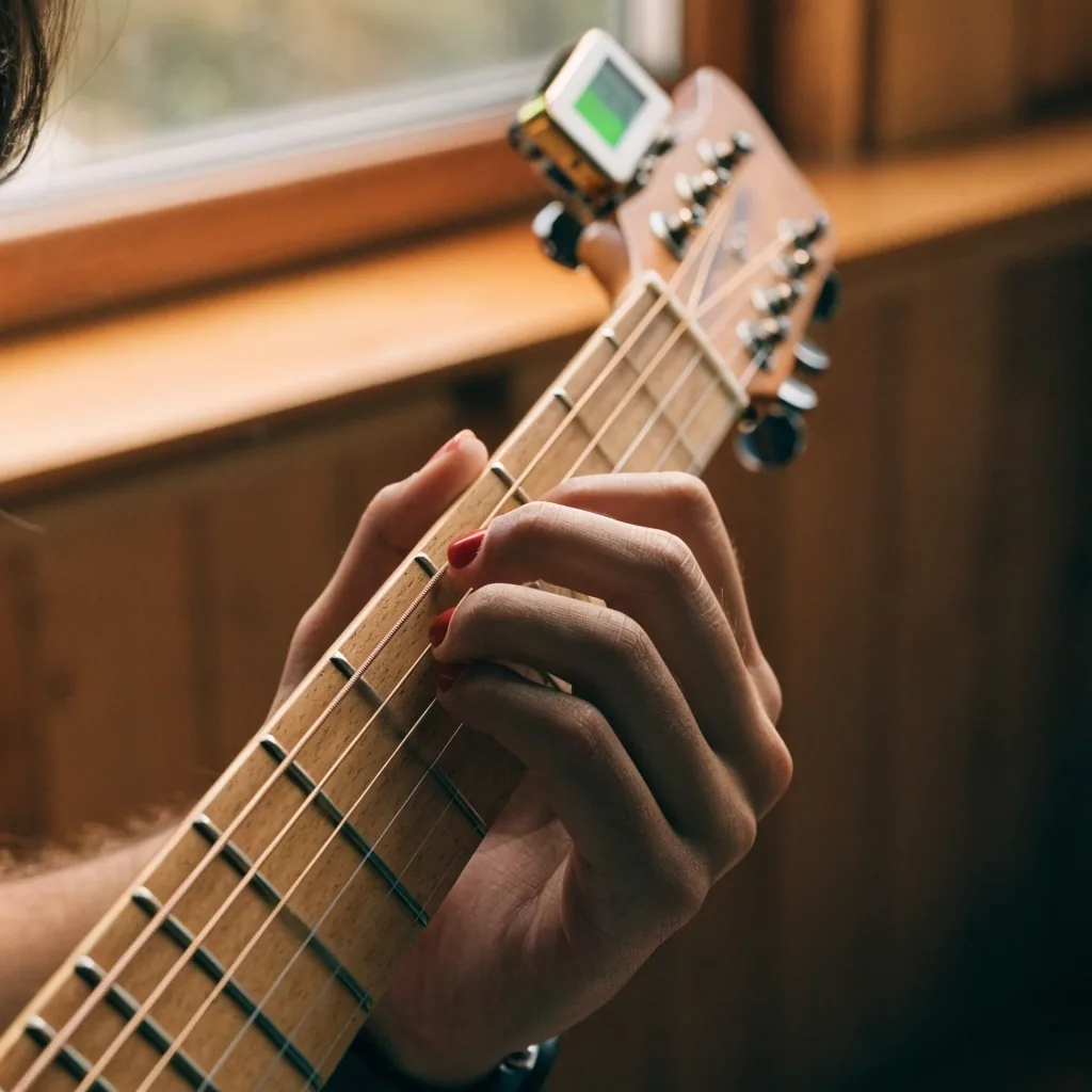 Close view of beginner fingers pressing chords on an acoustic guitar with a clip-on tuner on the headstock
