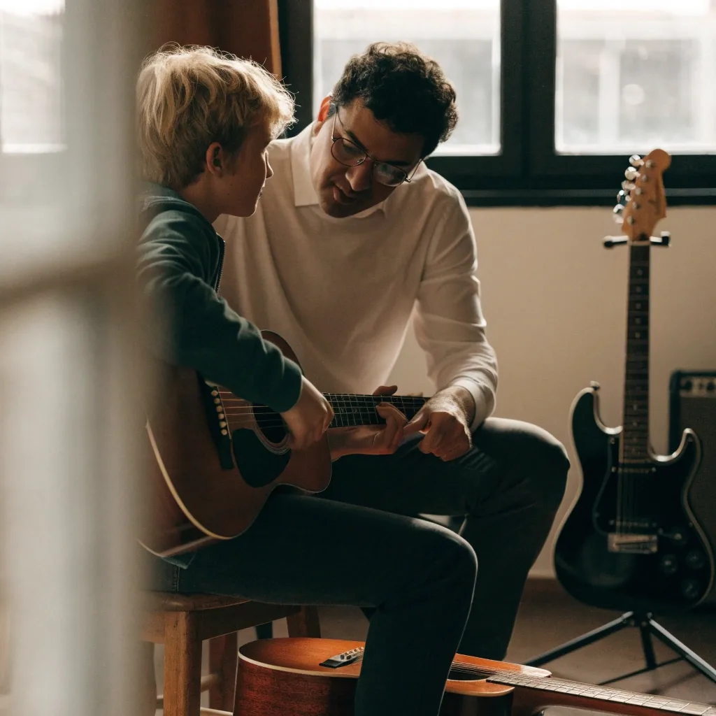 A teacher watching a learner's fretting hand during a one-to-one guitar lesson in a bright practice room