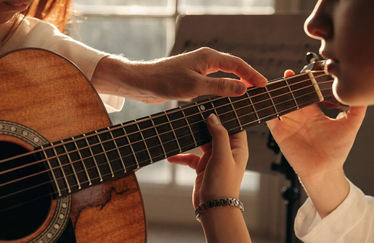 A teacher adjusting a learner's thumb position behind the guitar neck during a lesson